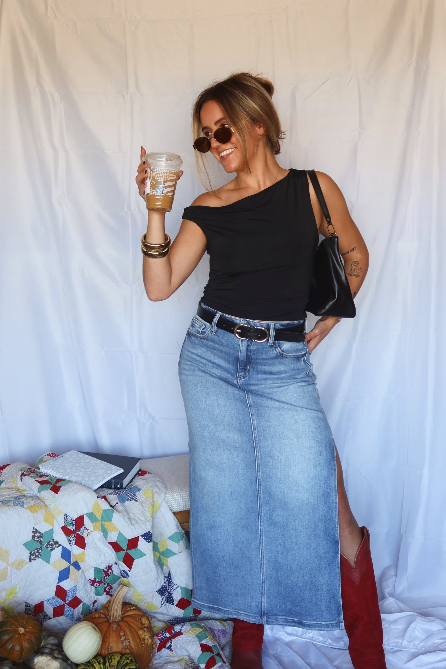 Woman in black top and blue denim skirt holding a drink against a white backdrop